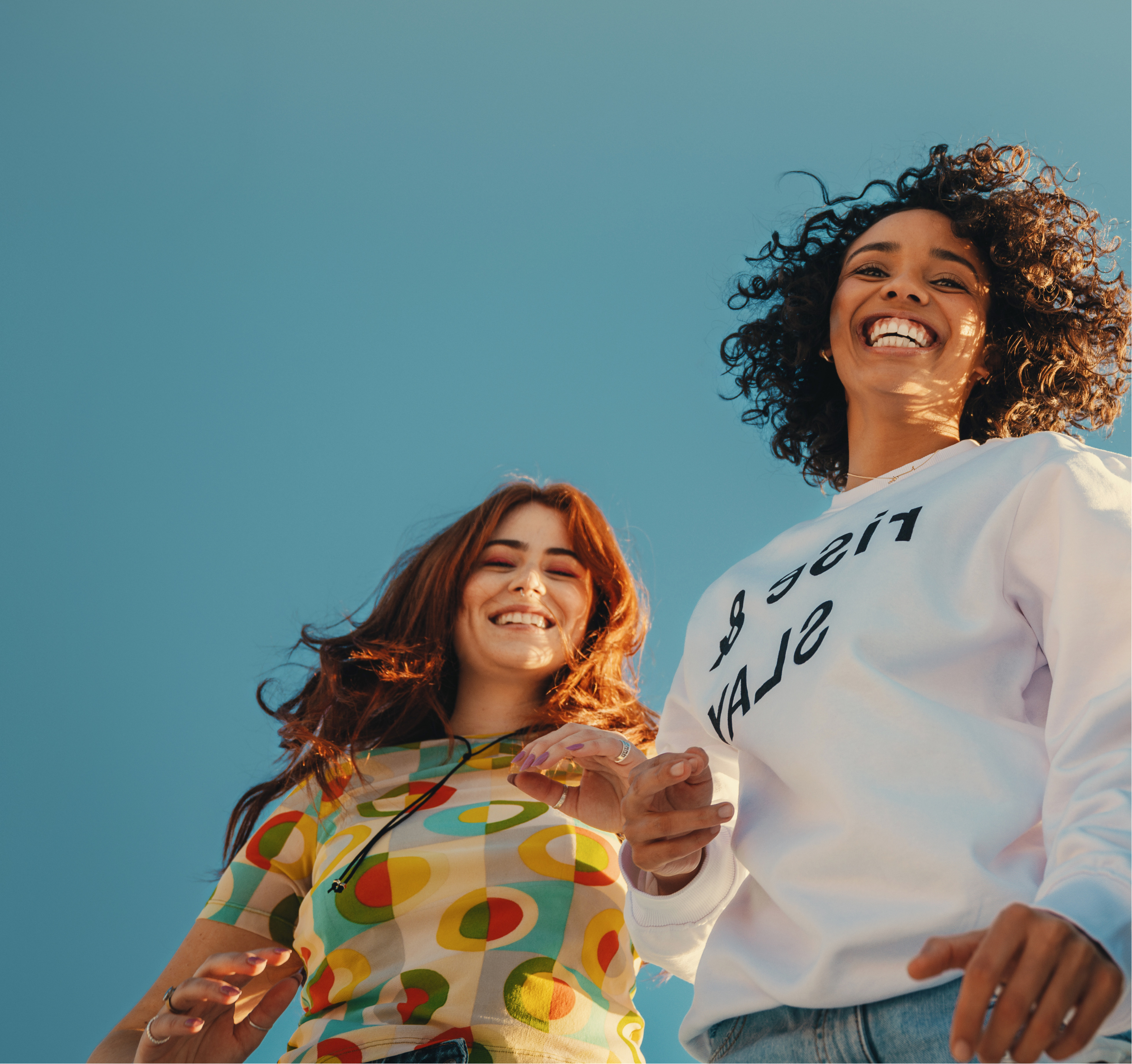 Two women standing against a clear blue sky, one wearing a colorful sweater and the other a white sweatshirt with text.